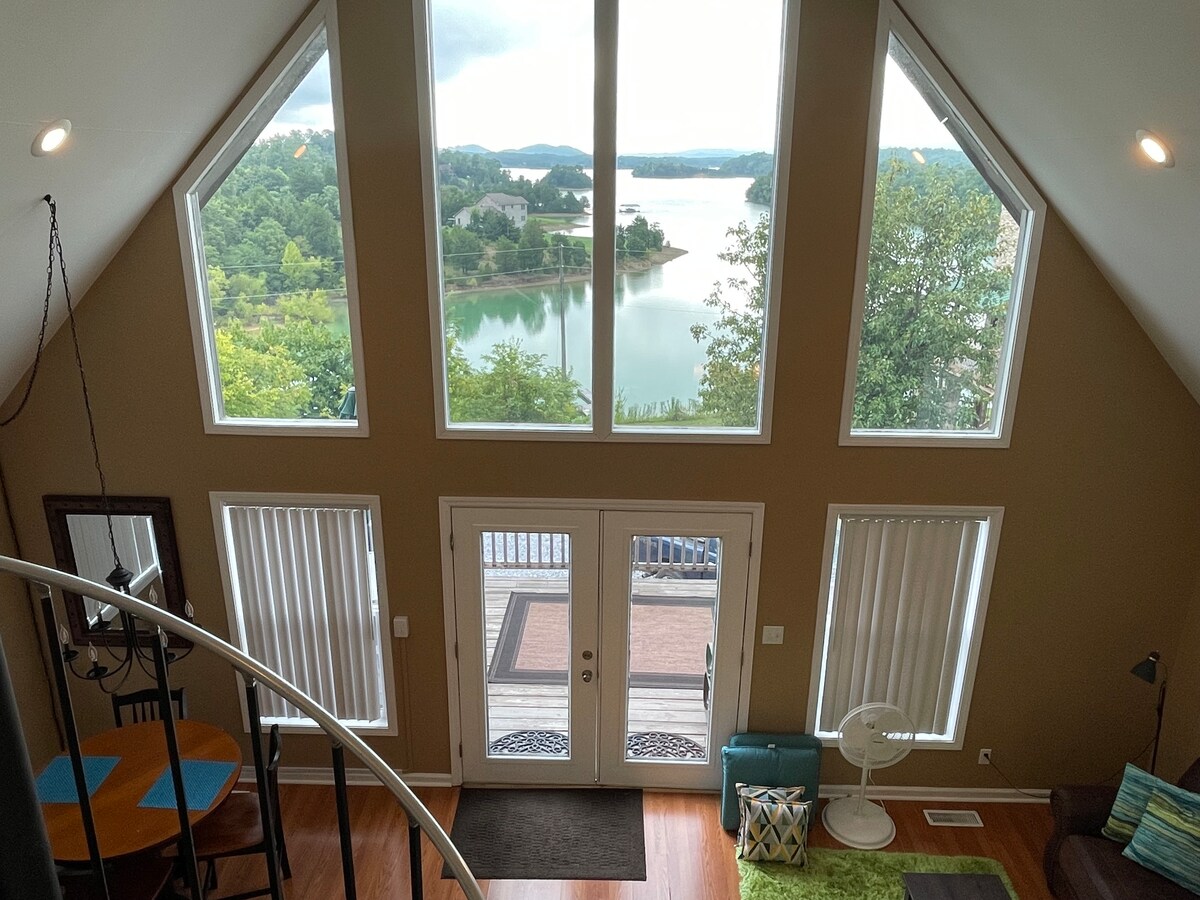 A view from above showcases tall, triangular windows framing serene lakeside scenery. Natural light fills the open space, illuminating a wooden dining area and inviting seating. The entrance features double doors, leading to a patio, with lush greenery visible outside.