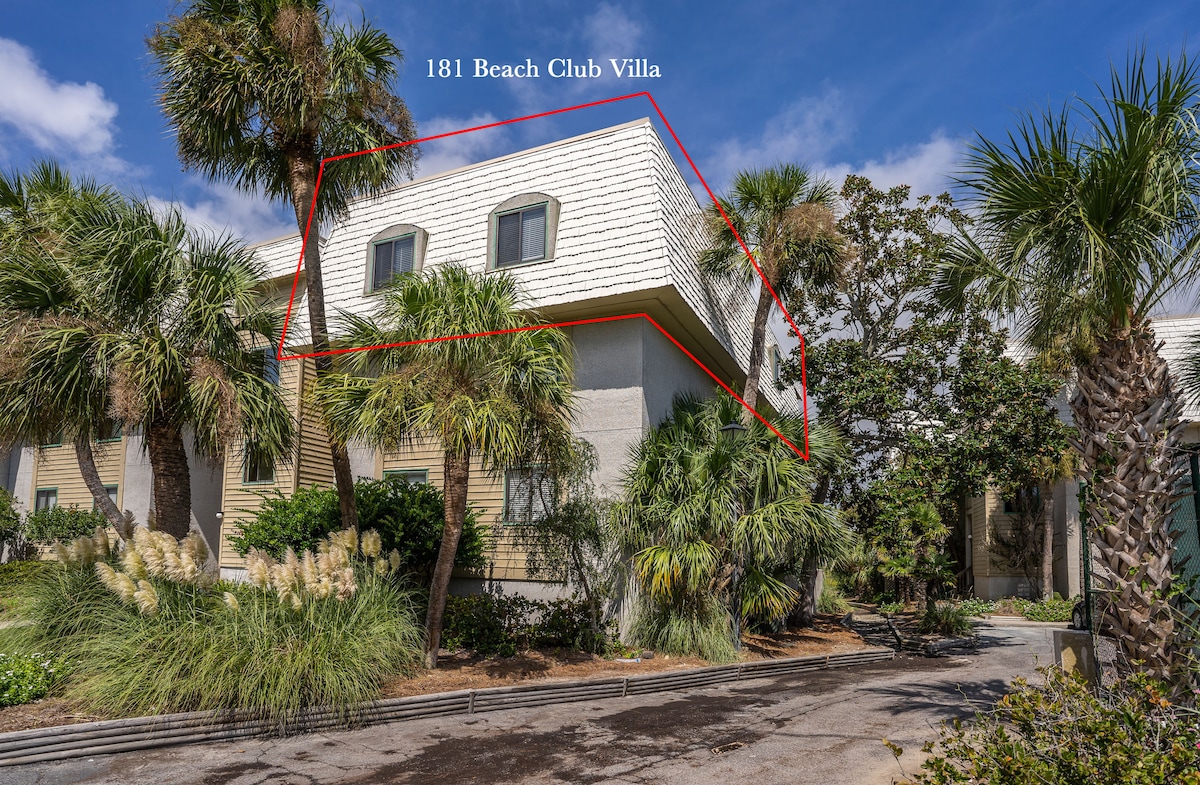 A corner unit of the Beach Club Villa is shown, featuring a light-colored exterior and a peaked roof. Lush palm trees and tropical vegetation surround the entrance. Large windows can be seen, reflecting the bright blue sky and providing an inviting ambiance.