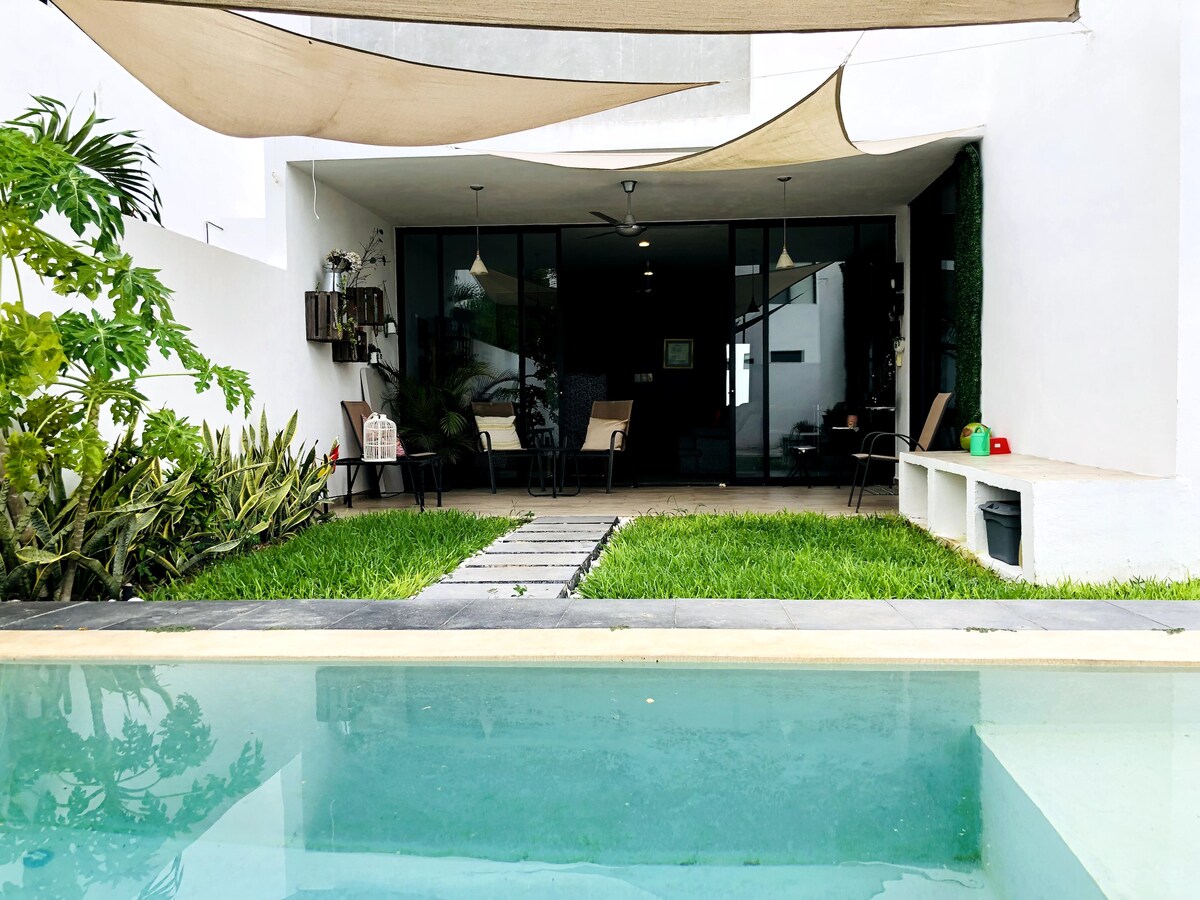 A tranquil outdoor space features a refreshing pool with clear water, surrounded by lush greenery. The area is shaded by fabric canopies, and a sleek patio is furnished with comfortable seating. Large glass doors provide a seamless transition to the interior.