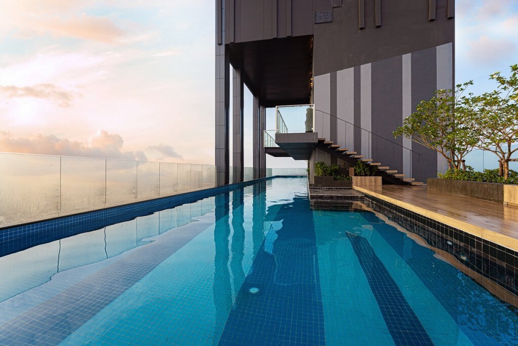 An elongated swimming pool is visible on the rooftop, reflecting the sky above. The pool is fringed by a wooden deck, with a set of stairs leading to the upper level. The surrounding area features lush greenery, contributing to a serene ambiance.