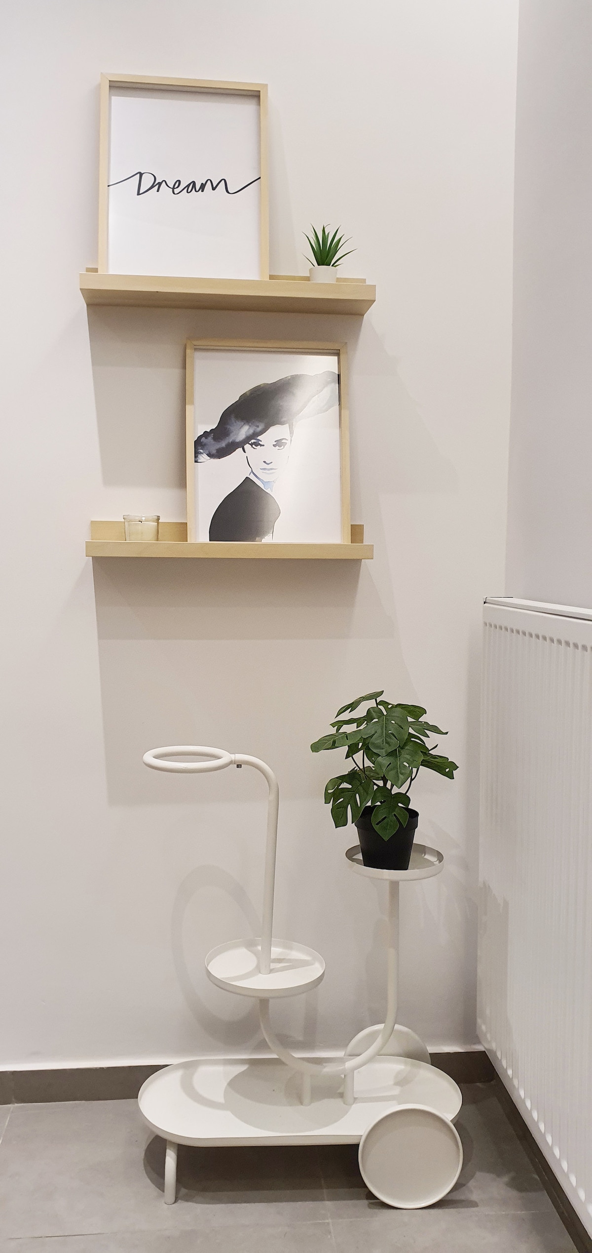 A wall corner showcases two wooden shelves holding a framed artwork and a motivational print. A small green plant is placed on a modern side table, complemented by a stylish white cart. The neutral wall enhances the contemporary decor.