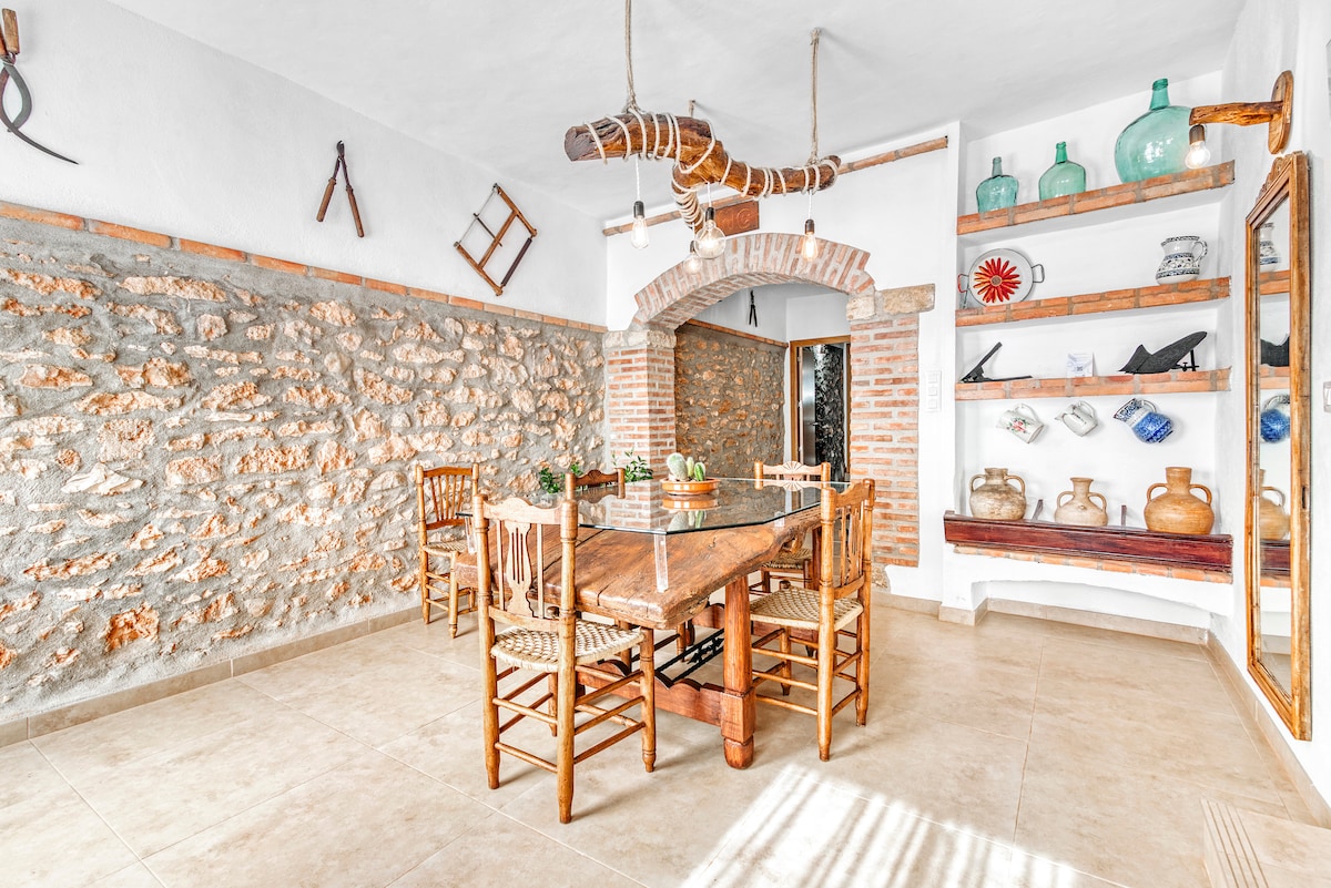 A spacious dining area features a long wooden table surrounded by chairs, with natural stone walls adding rustic charm. Decorative shelves display various colorful glass items and pottery, while a unique wooden beam overhead enhances the room's character.