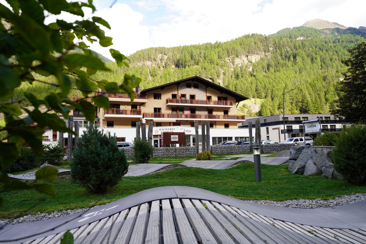 A multi-story building is set against a backdrop of green mountains, surrounded by manicured lawns and pathways. The structure features multiple balconies and large windows, allowing natural light to fill the spaces. Nearby greenery and rocky accents add to the tranquil outdoor environment.