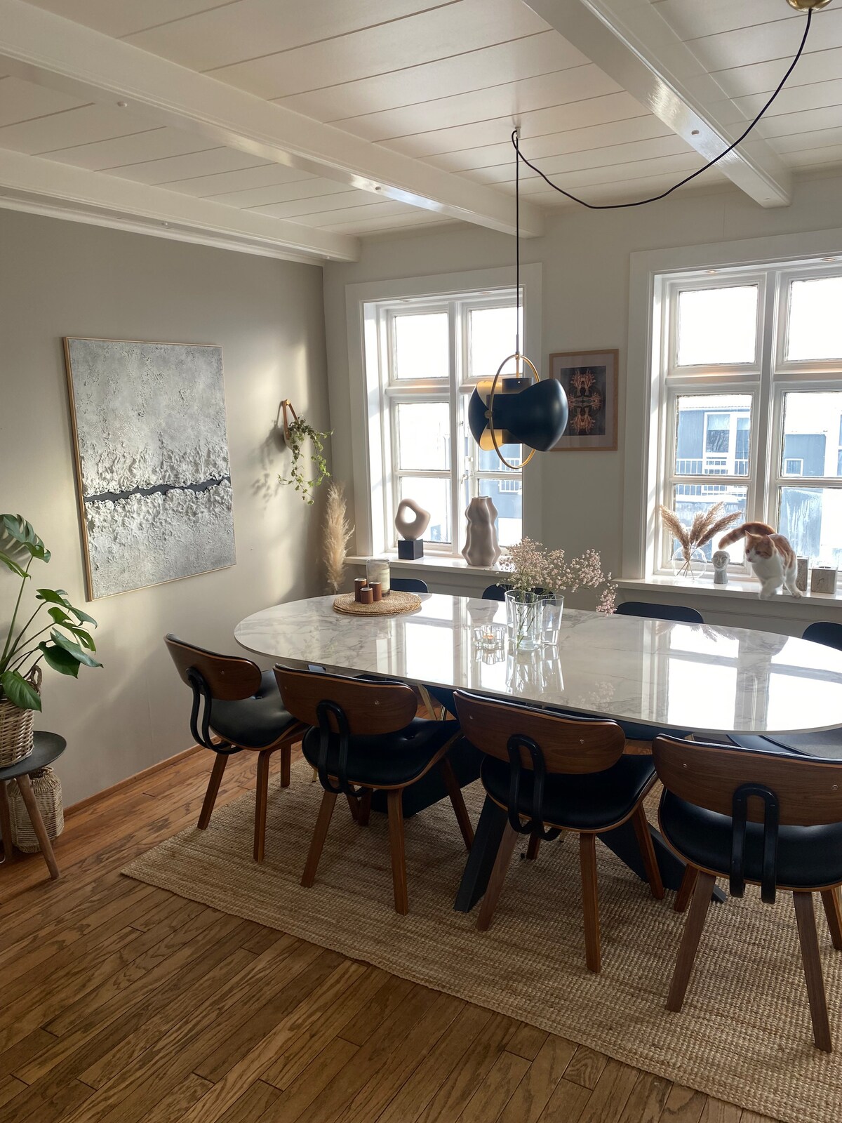 A spacious dining area features a large marble table surrounded by several wooden chairs. Natural light enters through multiple windows, illuminating a decorative wall art piece and various indoor plants. A soft area rug enhances the warm wooden flooring.
