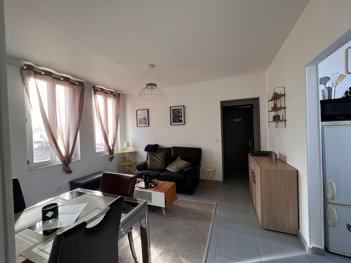 A modern living area features a dark sofa paired with a light wood coffee table and an accent chair. Large windows adorned with sheer curtains allow natural light. A dining table is set for four, and a small shelving unit is positioned against the wall.