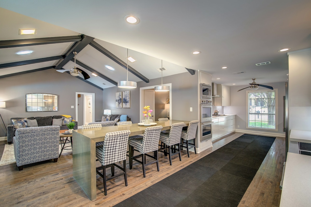 A modern open-concept kitchen and dining area features a long, light-colored dining table with ample seating. High-backed chairs with patterned upholstery complement the sleek kitchen appliances. Natural light enters through large windows, highlighting the vaulted ceilings and contemporary furnishings in this welcoming space.