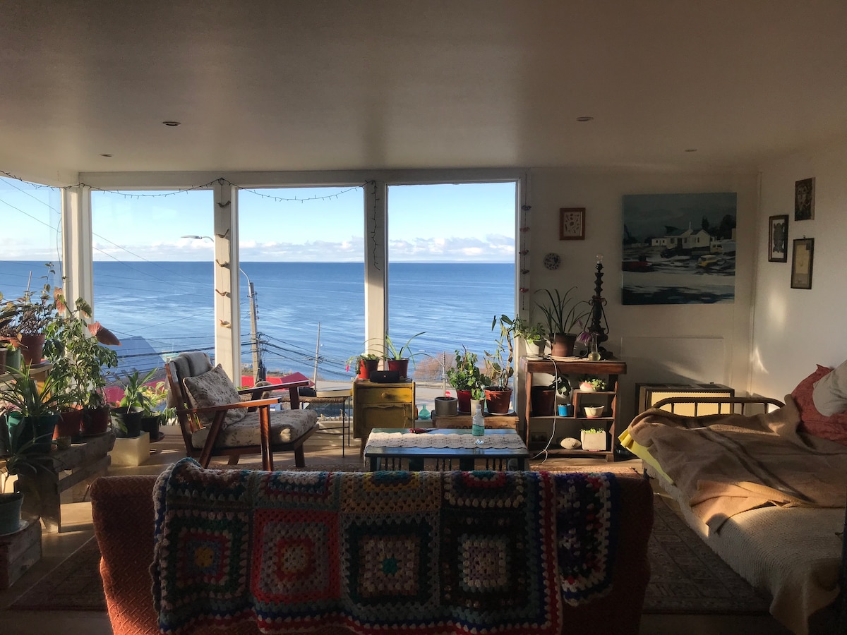 A bright living space showcases large windows with views of the sea, filled with natural light. Colorful plants adorn the room, including a variety of potted greenery. A cozy seating area features an armchair adjacent to a coffee table, with a patchwork blanket draped over a sofa.