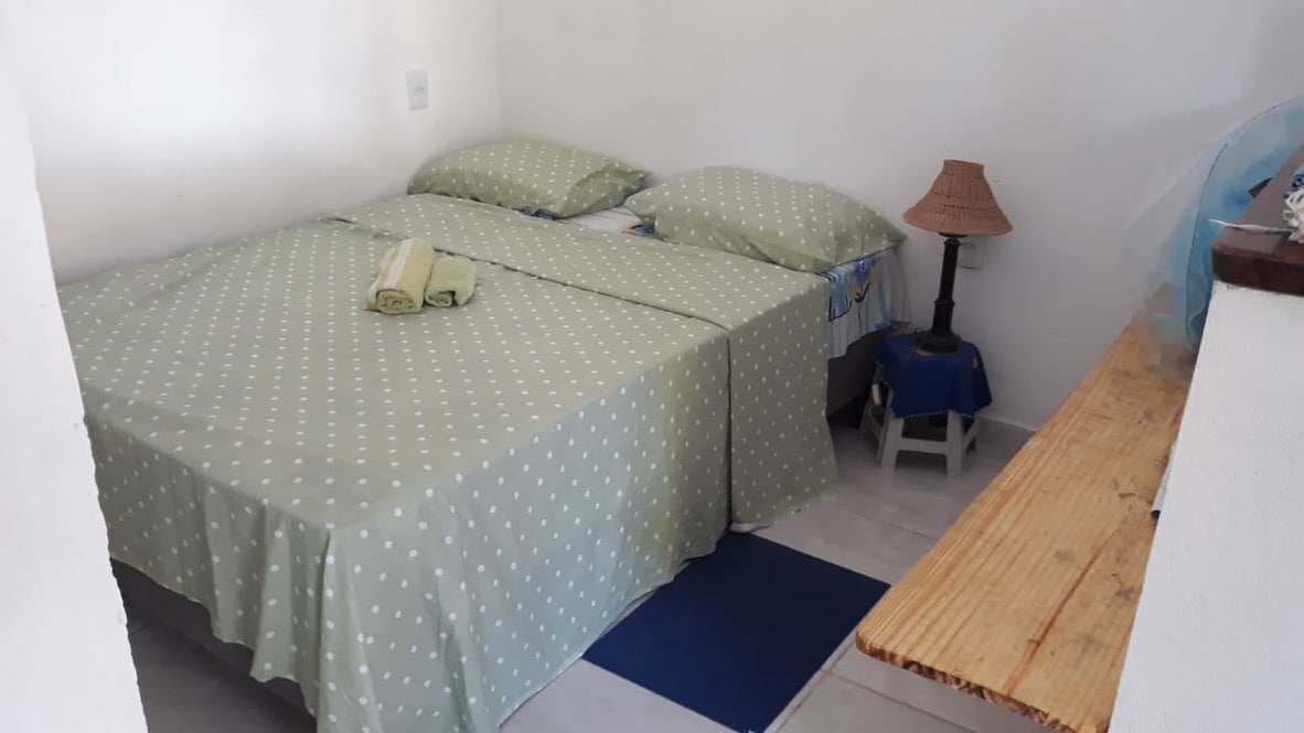 A cozy sleeping area is presented with a spacious bed covered in a light green polka dot coverlet. Two neatly arranged pillows rest at the head of the bed. A small bedside lamp and a wooden shelf add functionality to the simple white-walled room.