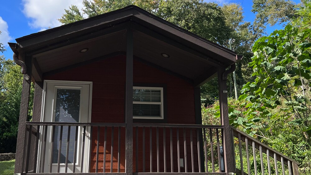 A charming cabin exterior is presented with a sloped roof and wooden porch. The entrance features a glass door alongside a small window. Lush green foliage surrounds the cabin, enhancing its natural setting against a bright blue sky with scattered clouds.