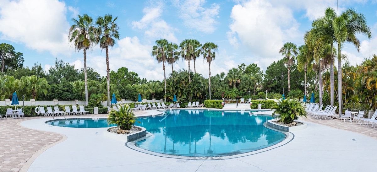 The expansive pool area is surrounded by lush palm trees, offering a relaxing atmosphere. Multiple lounge chairs are positioned around the water, and umbrellas provide shade. The clear blue waters reflect the sky, creating a tranquil setting for guests.