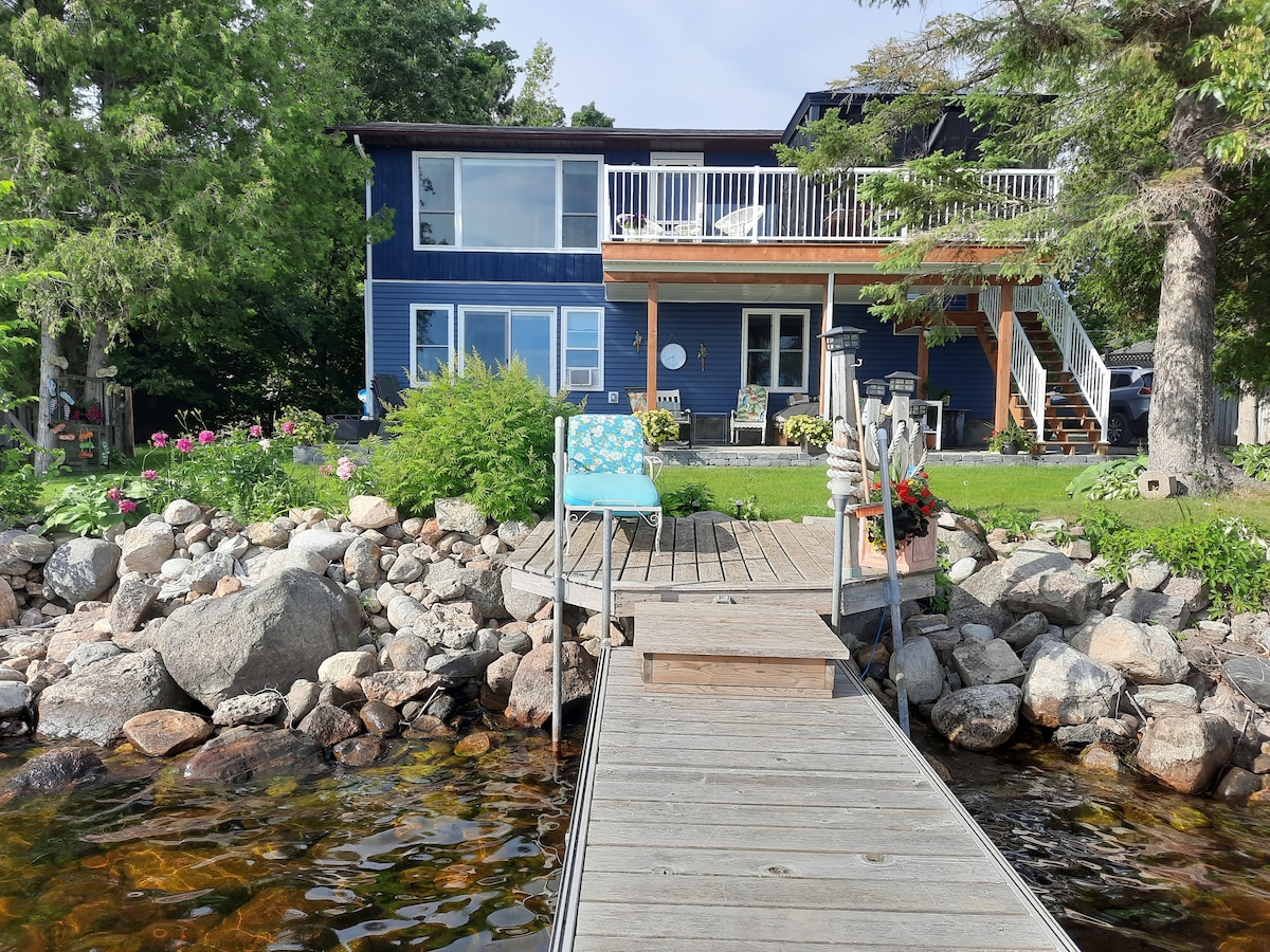 A wooden dock extends into the calm waters, leading to a blue and wooden two-story lakehouse. The house is surrounded by greenery and flower beds. A comfortable chair rests on the dock, inviting relaxation by the water. A private upper deck is visible above the entrance.
