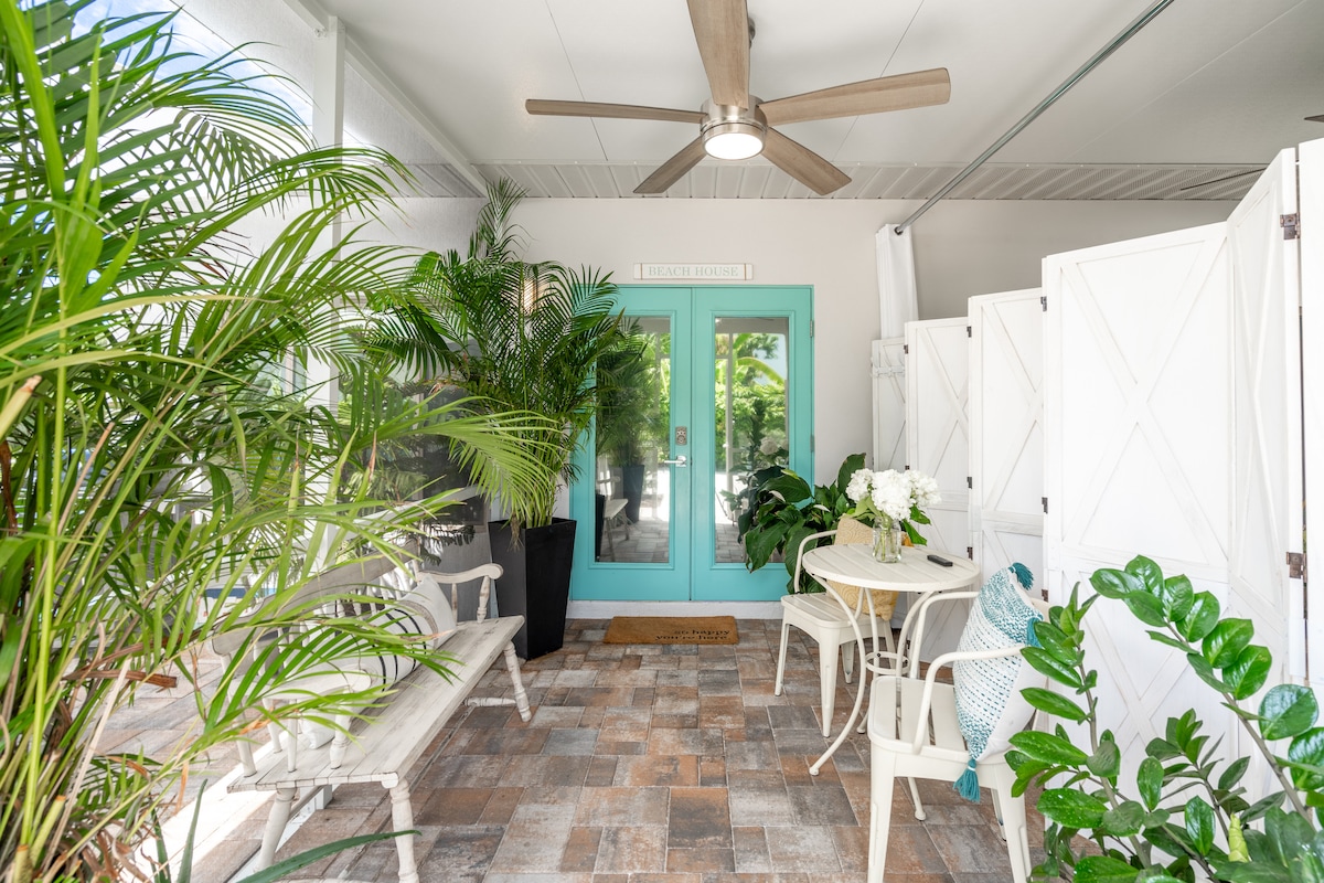 A covered patio space is surrounded by lush greenery and decorative plants. A light-colored table and two chairs are arranged on a tiled floor. Large, double doors in a vibrant blue lead to the outdoors, enhancing the welcoming atmosphere.