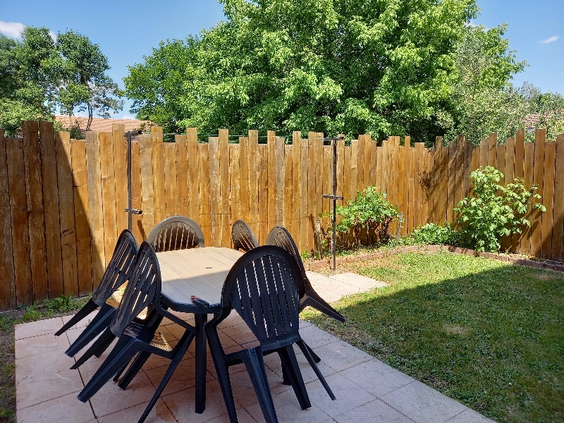 An outdoor patio area is featured, with a large wooden table surrounded by six black chairs. A wooden fence surrounds the space, providing privacy. Lush green plants are visible in the garden, along with a clear blue sky overhead.