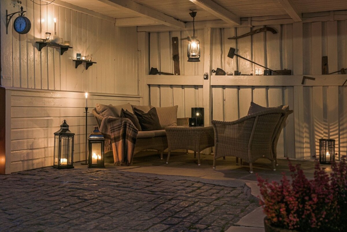 An outdoor seating area is framed by a wooden structure, featuring cozy wicker furniture arranged invitingly. Soft lighting from lanterns casts a warm glow, and decorative elements such as tools are displayed on the walls. A plaid blanket adds a touch of comfort.
