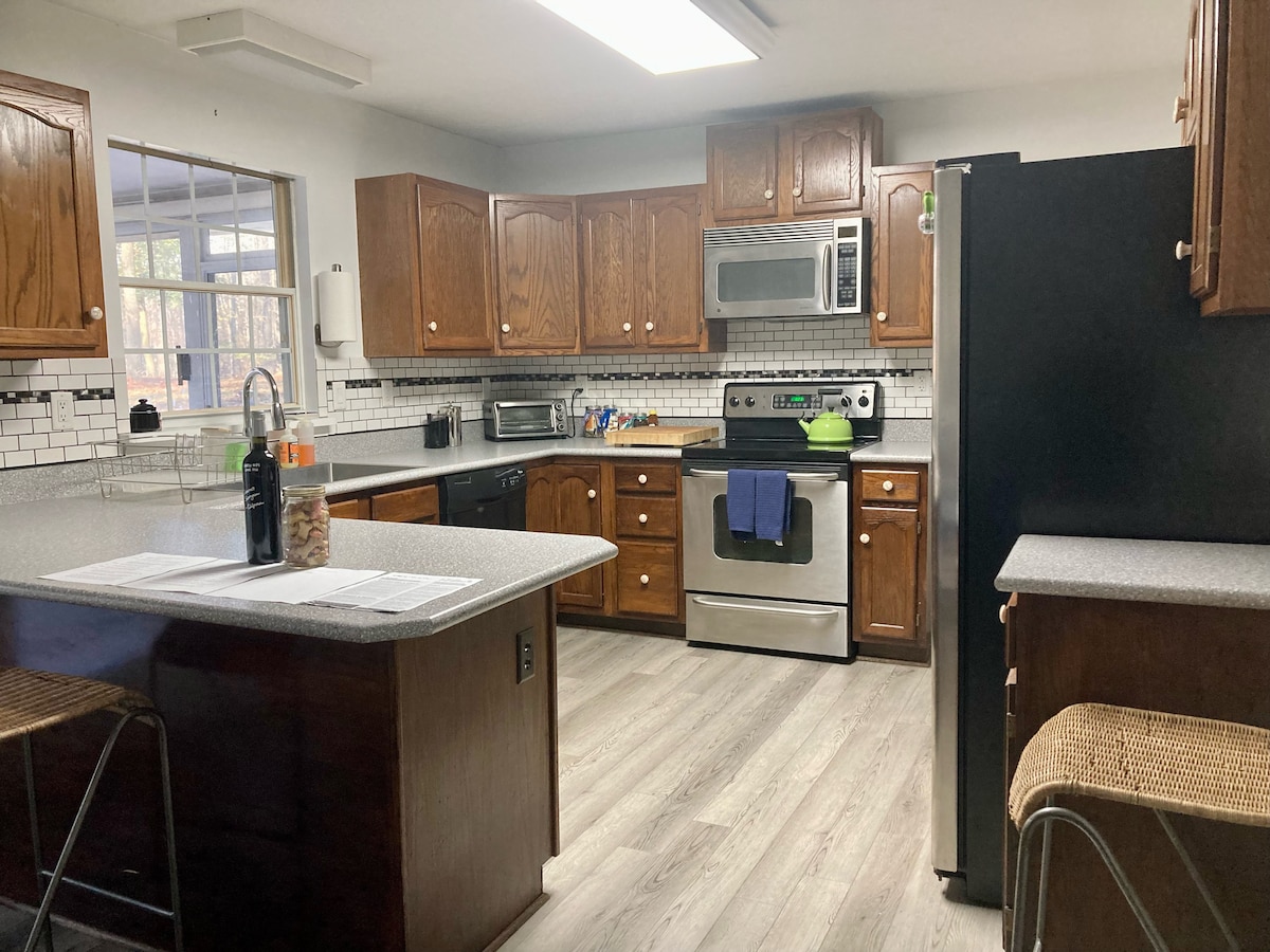 The kitchen features wooden cabinetry, modern appliances, and a spacious countertop. A stainless steel refrigerator and oven are complemented by a black dishwasher. Bar seating for two is available at the island, with natural light streaming in from the nearby window.