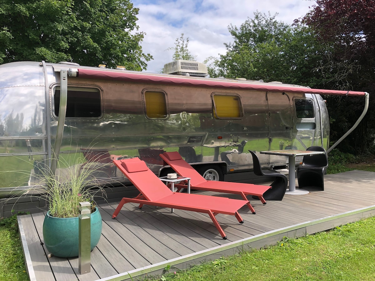 An Airstream caravan is positioned on a wooden deck, featuring a red awning. Red lounge chairs and a black table are arranged for relaxation. Lush greenery surrounds the space, enhancing the natural setting.