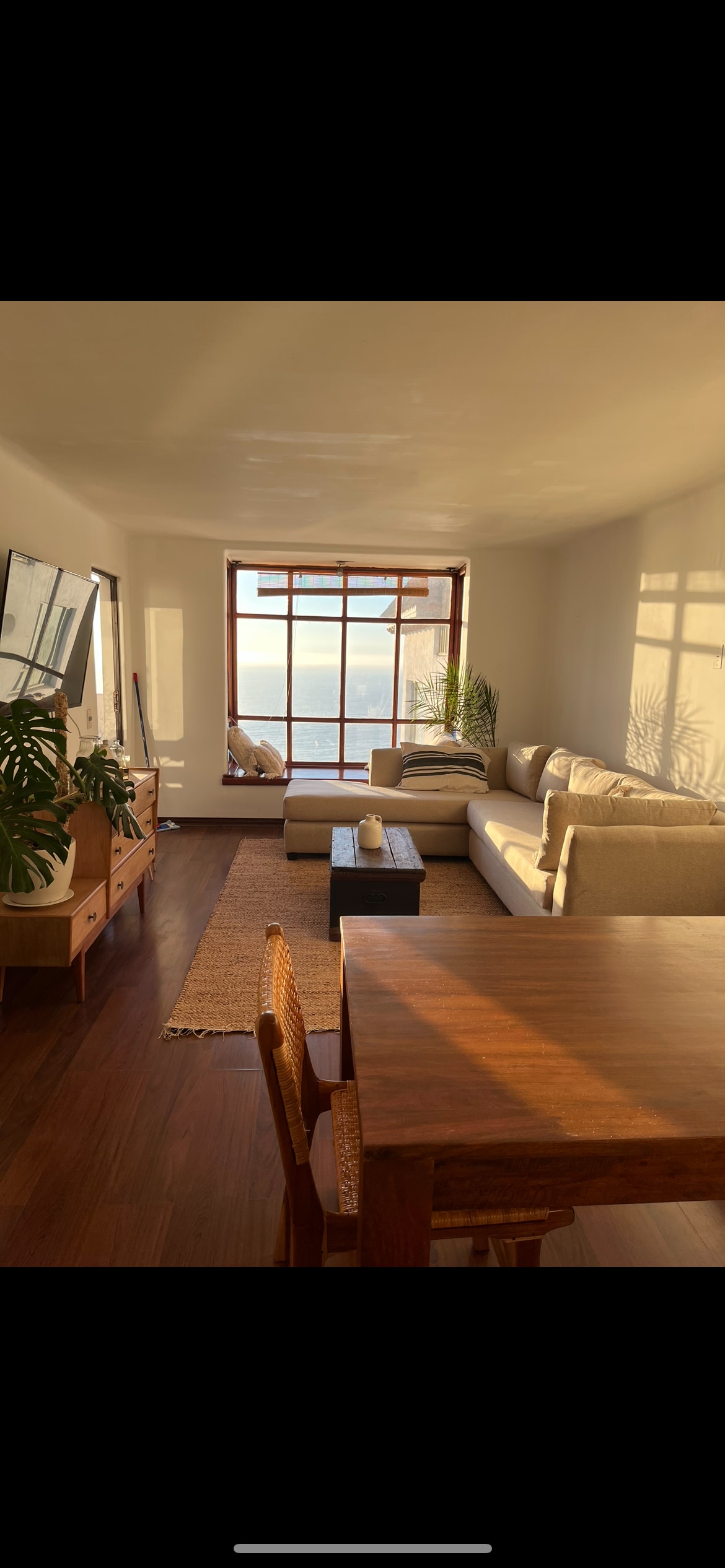 A bright living area features a large window overlooking the ocean, allowing natural light to fill the space. A comfortable sectional sofa and a low coffee table create a relaxed seating arrangement. A dining table is positioned nearby, complementing the wooden decor elements.