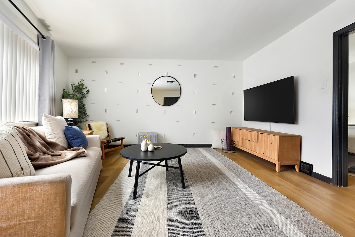 The living room features a light-colored sofa paired with a round black coffee table. A large wall-mounted TV is positioned opposite a minimalist media console. Natural light fills the space, highlighting a textured area rug and a circular mirror mounted on the wall.