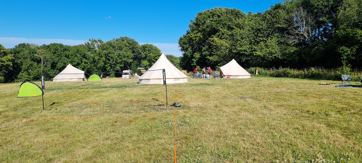 Spacious campsite featuring several bell tents arranged on open grassy land. Lush trees encircle the area, and outdoor games are positioned on the ground. A clear blue sky enhances the serene atmosphere of this eco-friendly camping experience.