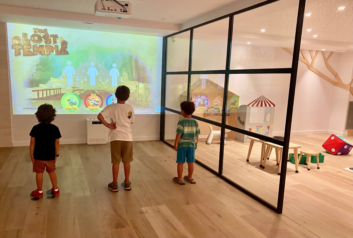 A playroom is highlighted, featuring children engaged with an interactive screen displaying a game. A glass partition separates the play area from additional toys and activities, all set against light wooden flooring and bright ambient lighting.
