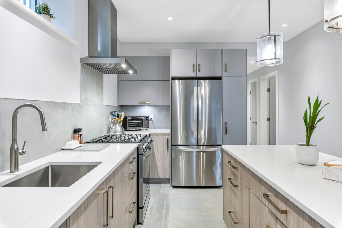 A modern kitchen features stainless steel appliances, including a gas range and a double-door refrigerator. Light wood cabinetry complements the white countertops. A spacious sink is positioned at one end, with a potted plant adding a touch of greenery on the countertop.
