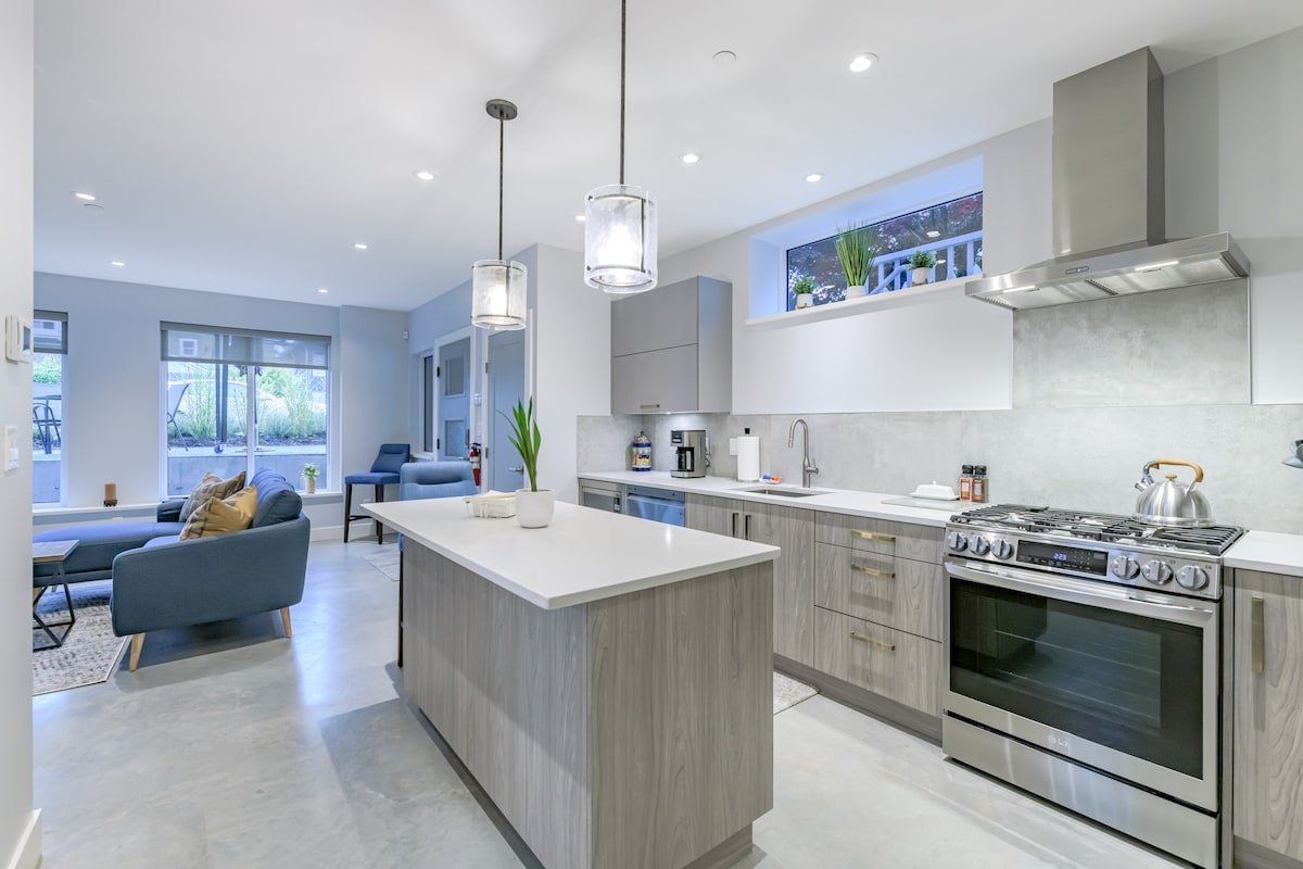 A modern kitchen features sleek cabinetry and a large central island with seating. Stainless steel appliances include a gas stove and coffee maker. Large windows allow natural light to illuminate the space, while an open layout connects the kitchen to the cozy living area.