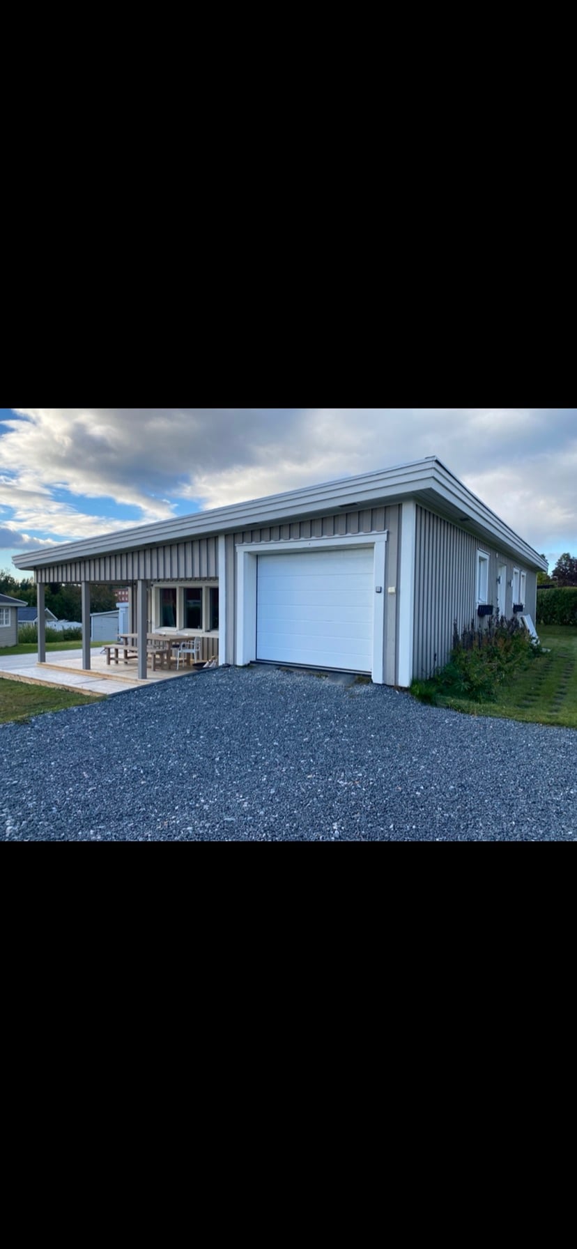 A modern, single-story building is presented with a light-colored exterior and a attached garage. A covered patio area features a seating arrangement facing the landscaped garden. The driveway is lined with gravel, and a grassy area extends beside the structure.