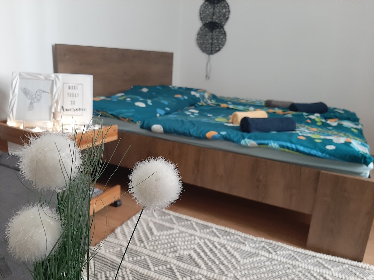 A spacious bed is dressed in a colorful bedding set, complemented by neatly arranged bath towels. A decorative framed quote is positioned on a nearby table, alongside a soft plant arrangement. The room features a simple wooden design and a patterned rug enhancing the overall decor.