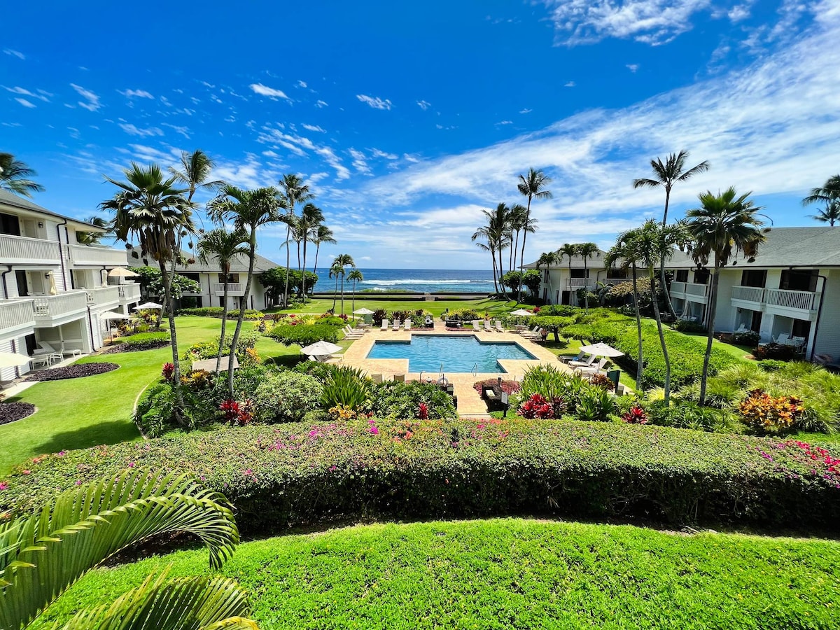 A scenic view showcases a lush garden with vibrant flowers and palm trees surrounding a central pool area. The sparkling water reflects the clear blue sky, while the vast Pacific Ocean is visible in the background, enhancing the inviting atmosphere of the resort.