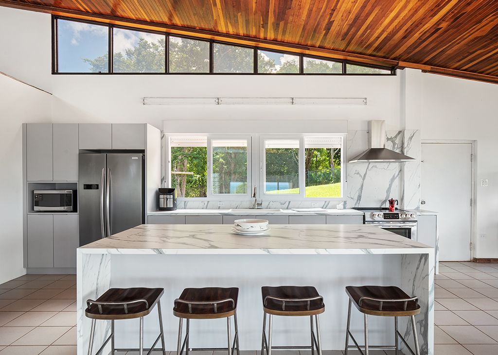 An open kitchen space features a large island with a white marble countertop and seating for four. Stainless steel appliances, including a refrigerator, microwave, and oven, are positioned against modern cabinetry. Natural light filters through large windows, illuminating the high wooden ceiling.
