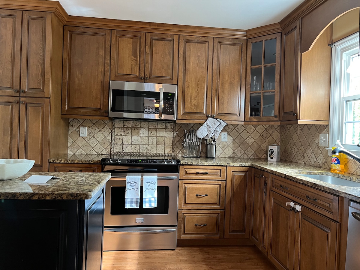 The kitchen features wooden cabinetry with a warm finish, complemented by granite countertops. A stainless steel microwave and oven are built into the cabinetry, while decorative tile backsplash enhances the space. Natural light enters through a window, illuminating the organized and functional cooking area.