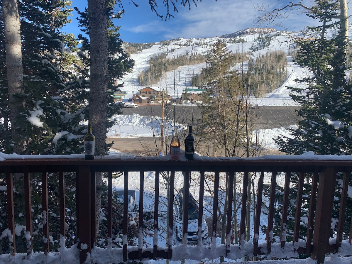 A scenic view from a wooden balcony showcases snow-covered slopes and tall trees. The landscape features a road leading to a lodge, with sunlight illuminating the serene winter surroundings. Two bottles are placed on the railing, adding a touch of detail to the scene.