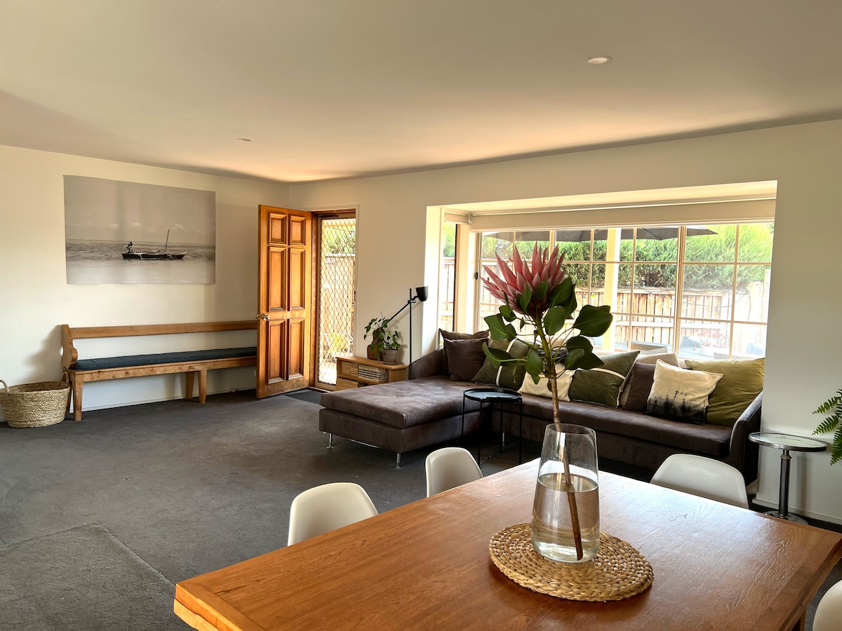 An open plan living area features a spacious couch positioned in a bay window, providing ample natural light. A wooden dining table occupies the foreground, complemented by contemporary chairs. A decorative plant and light decor add interest to the space, enhancing its welcoming feel.
