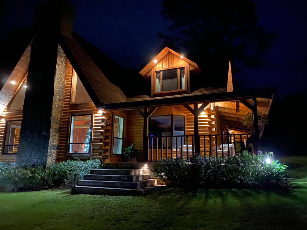 A charming log cabin is illuminated at night, showcasing wooden beams and stone accents. Warm light spills from the windows, and steps lead up to a spacious porch surrounded by well-maintained greenery. The inviting entrance reflects the cozy atmosphere of the property.