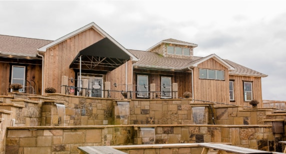 A rustic wooden structure is showcased with a wide covered entrance. The building features large windows and a series of stone steps leading up to a spacious patio area, surrounded by natural stone walls. The setting provides a distinctive charm.