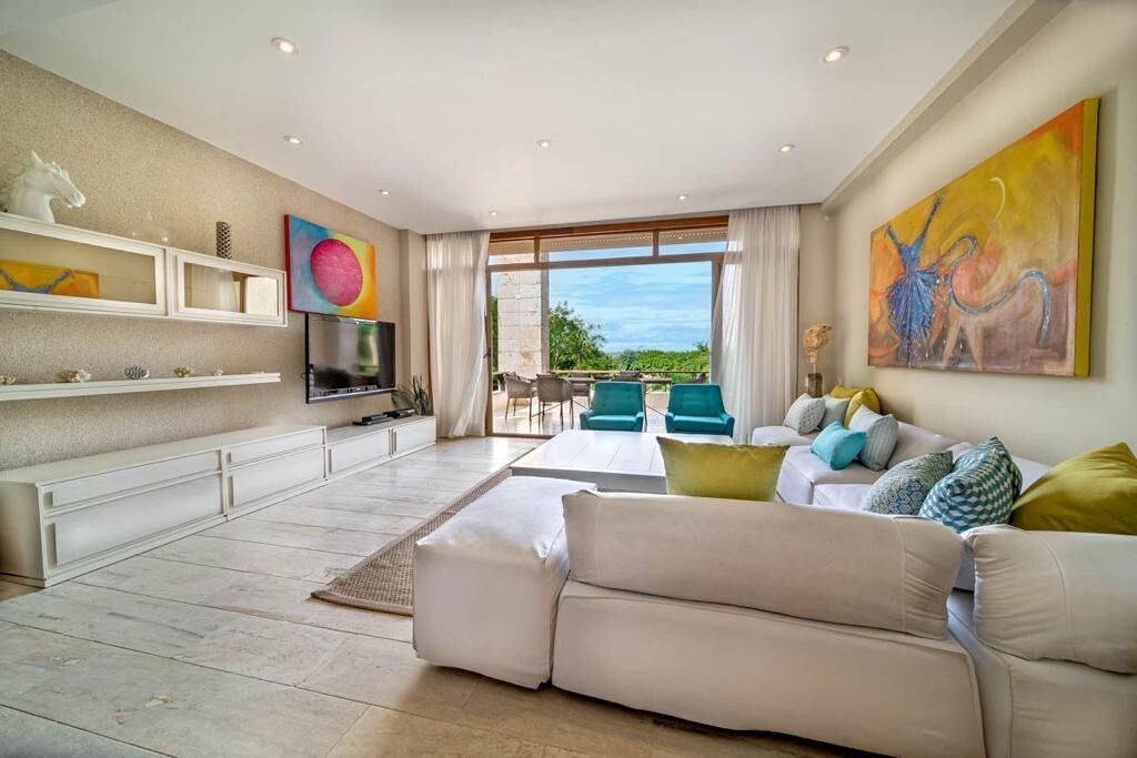 A bright living area features a comfortably arranged white sectional sofa with colorful pillows. An entertainment unit is mounted on the wall, and large sliding glass doors lead to an outdoor space with seating. Two armchairs provide additional seating, while vibrant artwork adorns the walls.