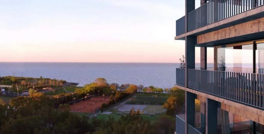 A view from the balcony showcases the serene waters of the river, framed by lush greenery and recreational facilities below. The horizon glows with soft pastel colors as the sun sets, creating a tranquil backdrop for the modern architecture of the complex.