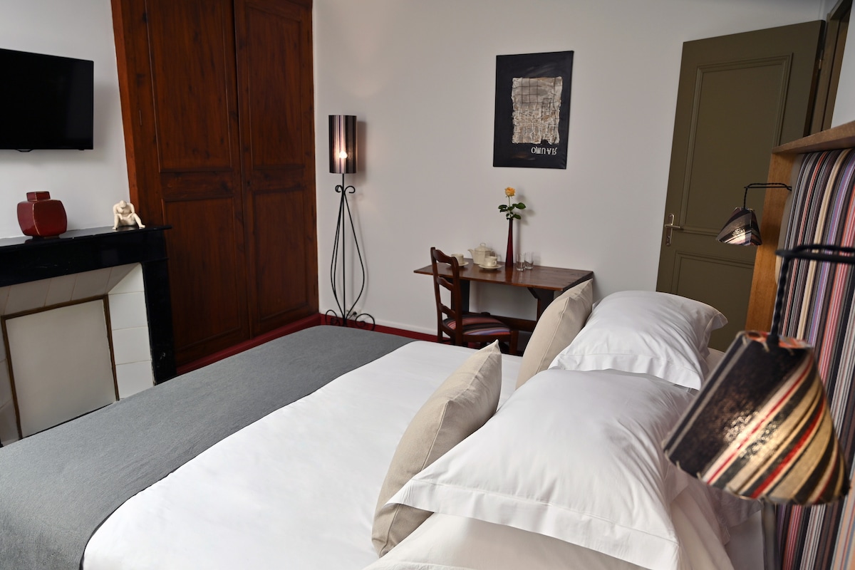 A spacious bedroom is shown with a large bed featuring plush pillows and a comfortable duvet. A dark wooden wardrobe and a small desk with a chair are visible, alongside a lamp providing gentle lighting. A framed artwork adorns the wall, complemented by a television mounted above the fireplace.