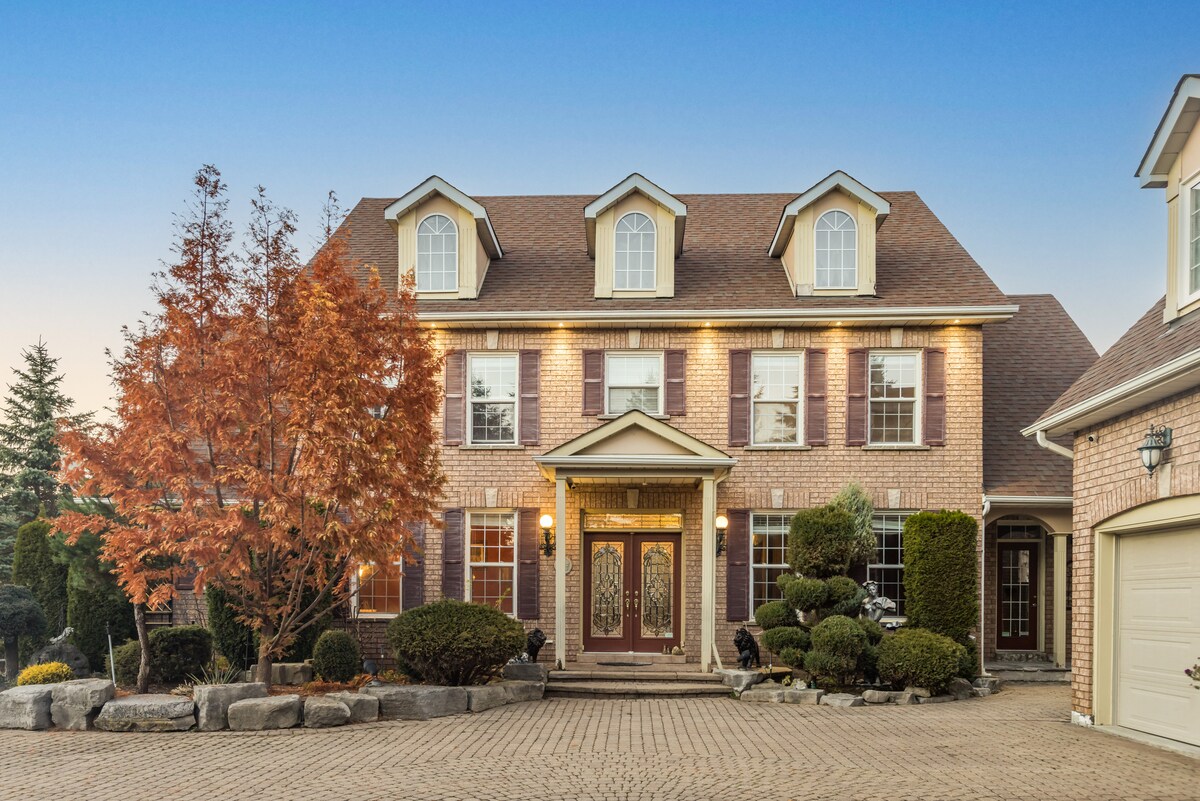 A stately brick home features a welcoming entrance with double doors and elegant lighting. Large windows reflect the landscape, while manicured shrubs and a vibrant tree offer seasonal color. A textured stone path leads to the front, enhancing the home's inviting setting.