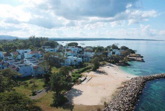 A serene beachfront area is visible with soft sandy shores leading to clear blue waters. Surrounding the beach, a collection of residential buildings and green trees is seen, set against a backdrop of distant hills and a softly clouded sky.