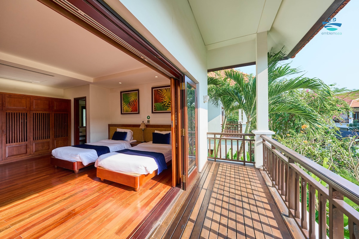 A spacious bedroom is featured with two single beds, complemented by natural light flowing through wide glass doors. A wooden balcony overlooks lush greenery, enhancing the serene atmosphere. The floors are crafted from polished wood, contributing to a warm and inviting feel.