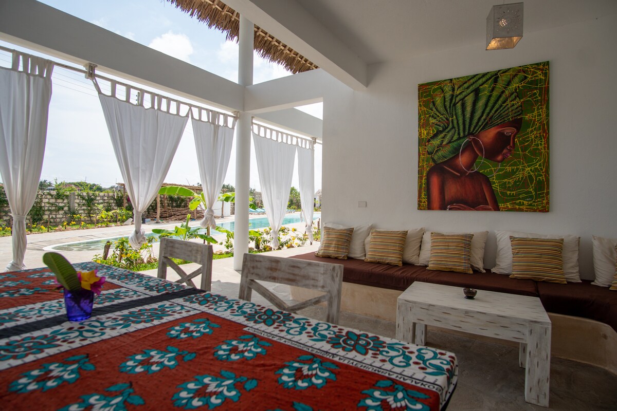 A spacious outdoor living area is presented, featuring a large table adorned with a colorful tablecloth. Comfortable seating with cushions is arranged along the wall. Sunlight filters through sheer curtains, while a vibrant piece of wall art adds a touch of character to the bright space.