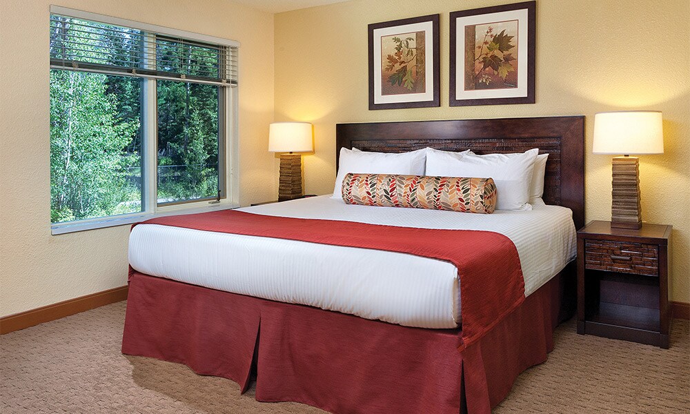 A spacious bedroom presents a king bed with a decorative red skirt, complemented by a patterned bolster pillow. Two bedside lamps illuminate the room, while nature views can be seen through the large window adorned with neutral-toned curtains. Framed wall art adds a touch of color.
