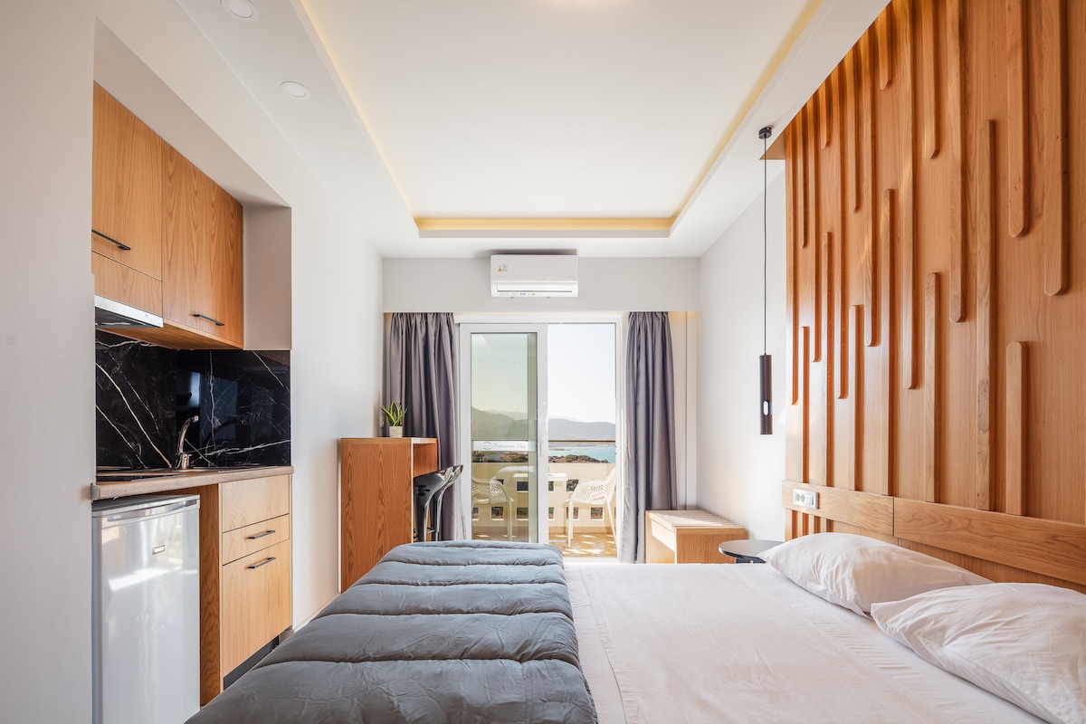 A modern room features a spacious bed with a textured wooden wall behind it. A kitchenette with wooden cabinetry is visible, alongside a small refrigerator. Natural light enters through sliding glass doors, leading to a balcony with outdoor seating and scenic views.