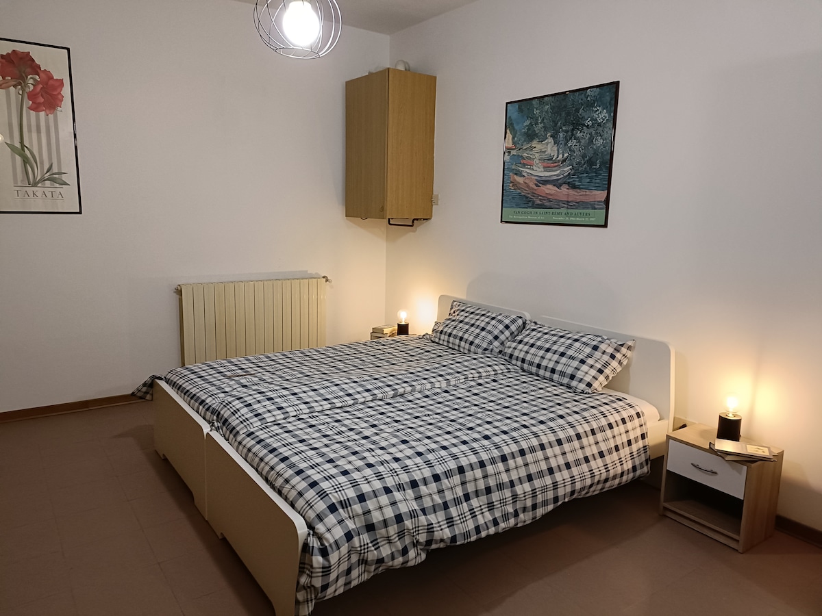 The sleeping area features a spacious bed with a checkered duvet cover. Nightstands with lamps are positioned on either side. A simple artwork adorns the wall, while a radiator provides warmth. Soft lighting enhances the welcoming ambiance of the room.