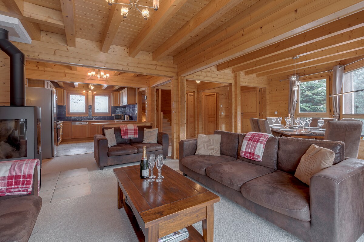 A warm living area is equipped with comfortable sofas and a wooden coffee table, creating a cozy gathering space. The room is characterized by wooden beams overhead and large windows allowing natural light to illuminate the space, revealing a glimpse of the forested surroundings.