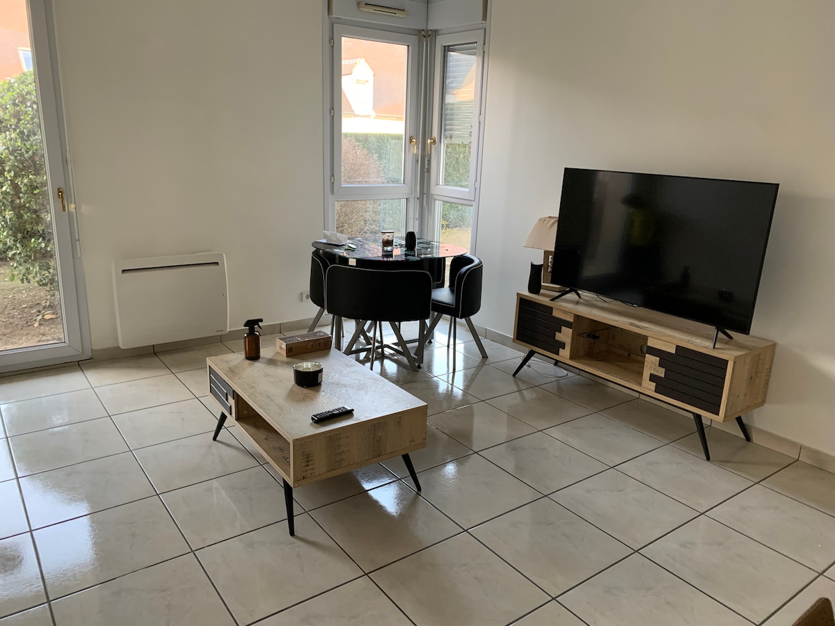 The bright living area features large windows allowing natural light. A television is positioned on a wooden media console, and a coffee table with a minimalist design is centered within the space. A dining table is set for four, enhancing the room's functionality.