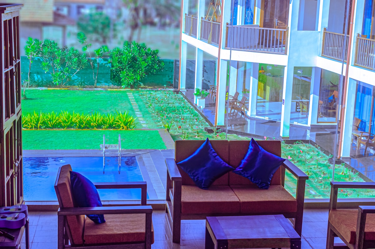 A shaded seating area features comfortable sofas adorned with purple cushions. A view of the lush green landscape is visible through large glass doors, revealing a swimming pool and well-maintained garden outside.
