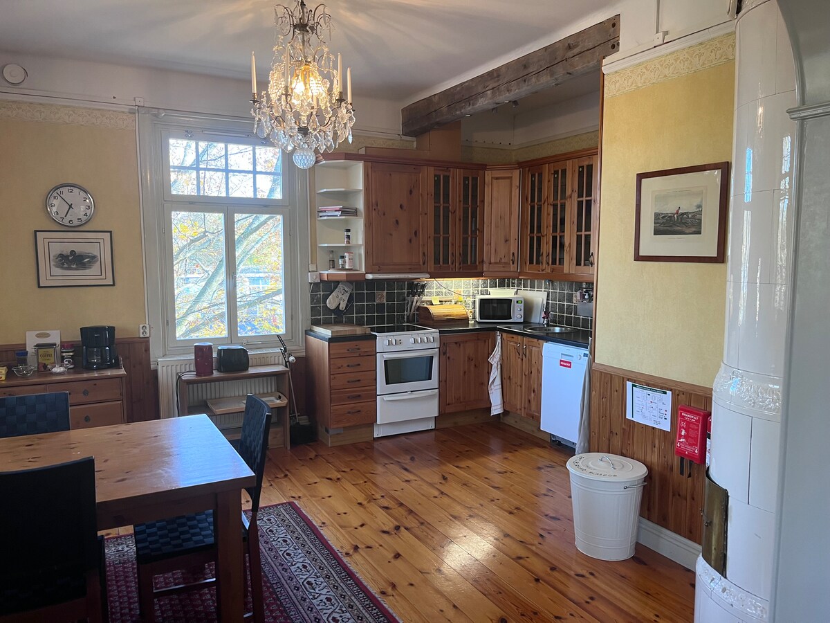 A well-equipped kitchen features wooden cabinetry and modern appliances. A dining table with four chairs is positioned near a window, allowing natural light to illuminate the space. Decorative elements include a chandelier and framed pictures on the walls, adding character to the room.