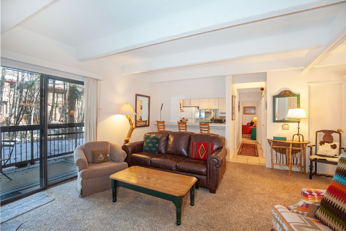 A welcoming living area is featured, showcasing a brown leather sofa and a light-colored armchair positioned around a wooden coffee table. Large sliding glass doors open to a patio, allowing natural light to brighten the space. Light carpeting and neutral walls create a comfortable atmosphere.